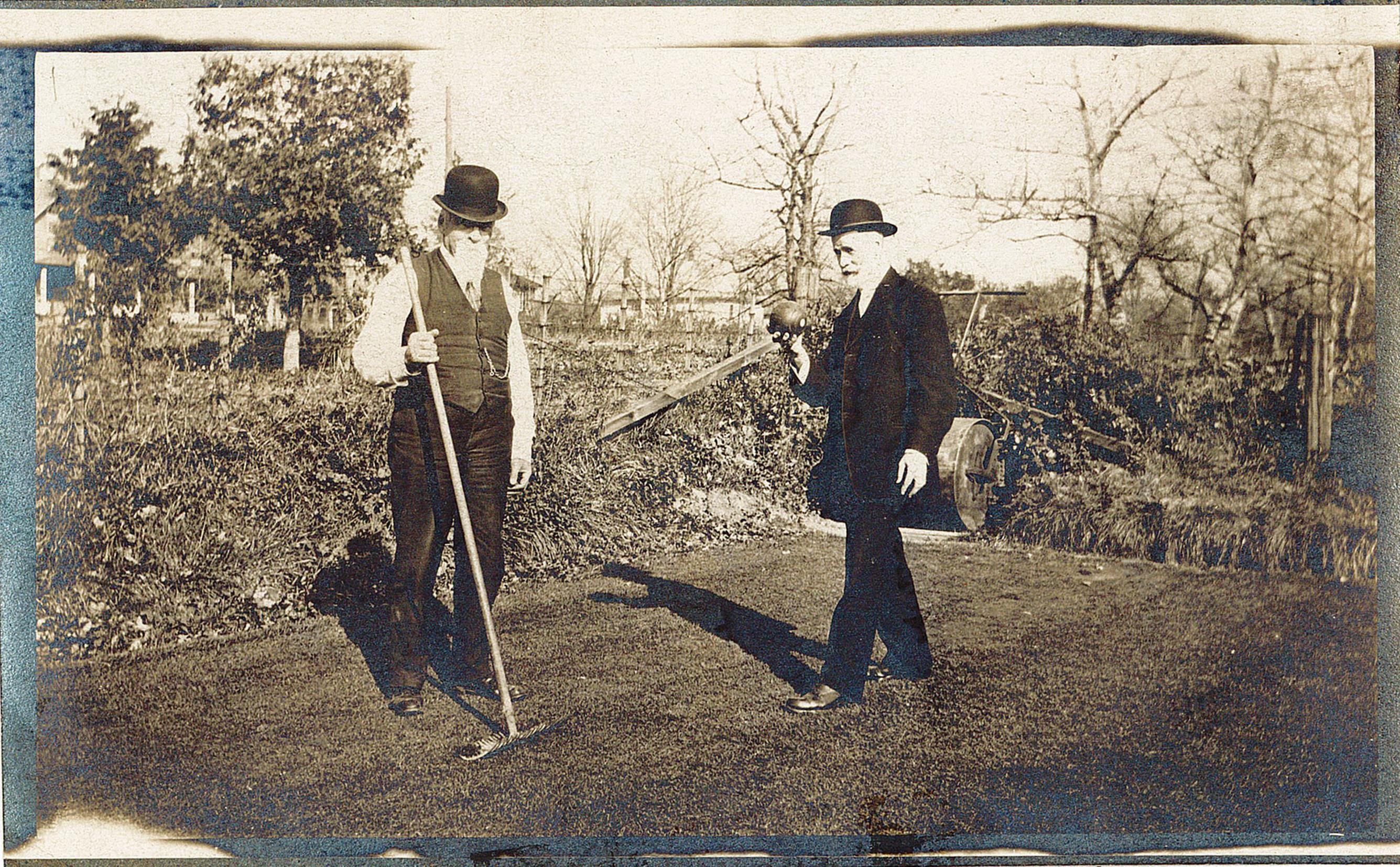 Photograph of two men playing lawn bowling. The man on the left is wearing a bowler hat and is dressed with a shirt, vest, and pants. He is holding a rake. The second man is holding a lawn bowling ball and is ready to roll the ball. He is also wearing a bowler hat and a suit.