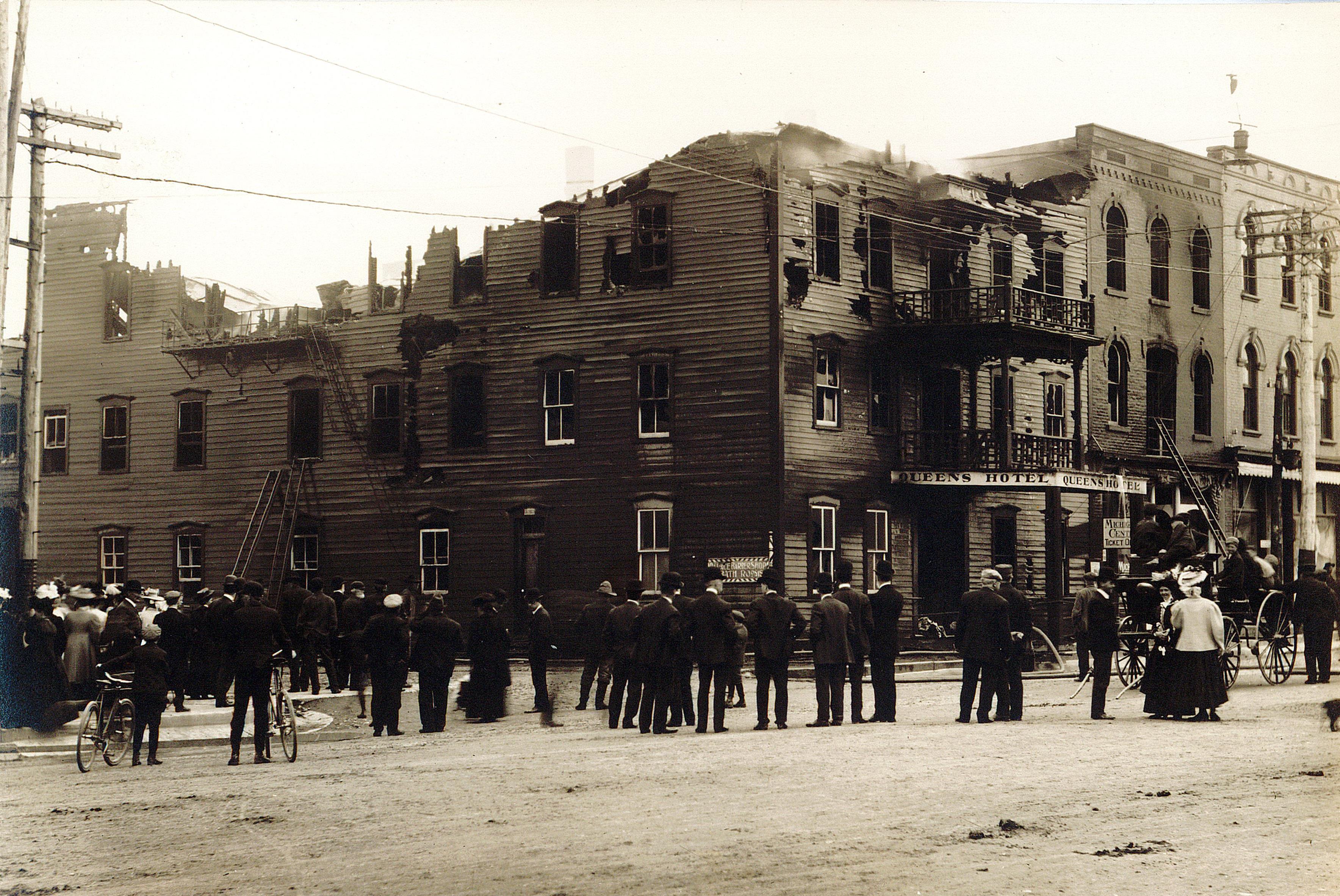 Photo of Queen's Hotel after a fire, top of building is missing. Smoke is escaping the interior of the building. A crowd observes the building from the street.