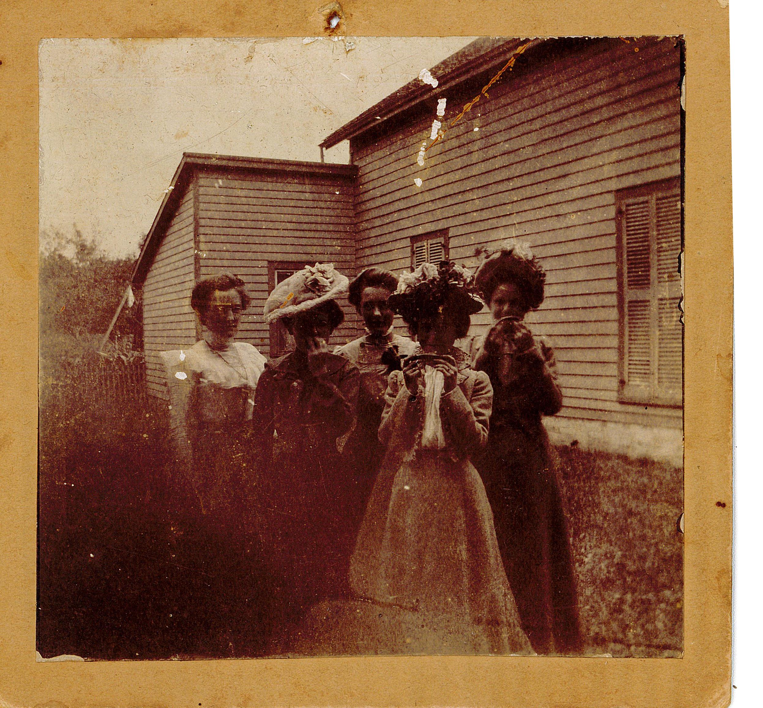 Five ladies posing in front of a wood frame building, wearing long dresses and fancy hats.