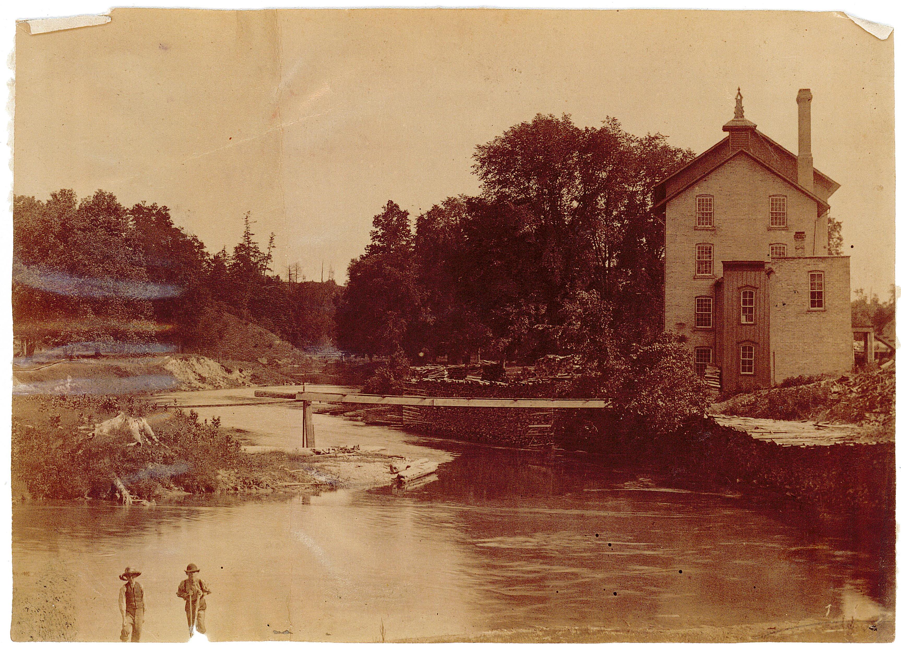 A view of E.D. Tillson's pea and barley mill, a four-storey brick building on a creek surrounded by trees.