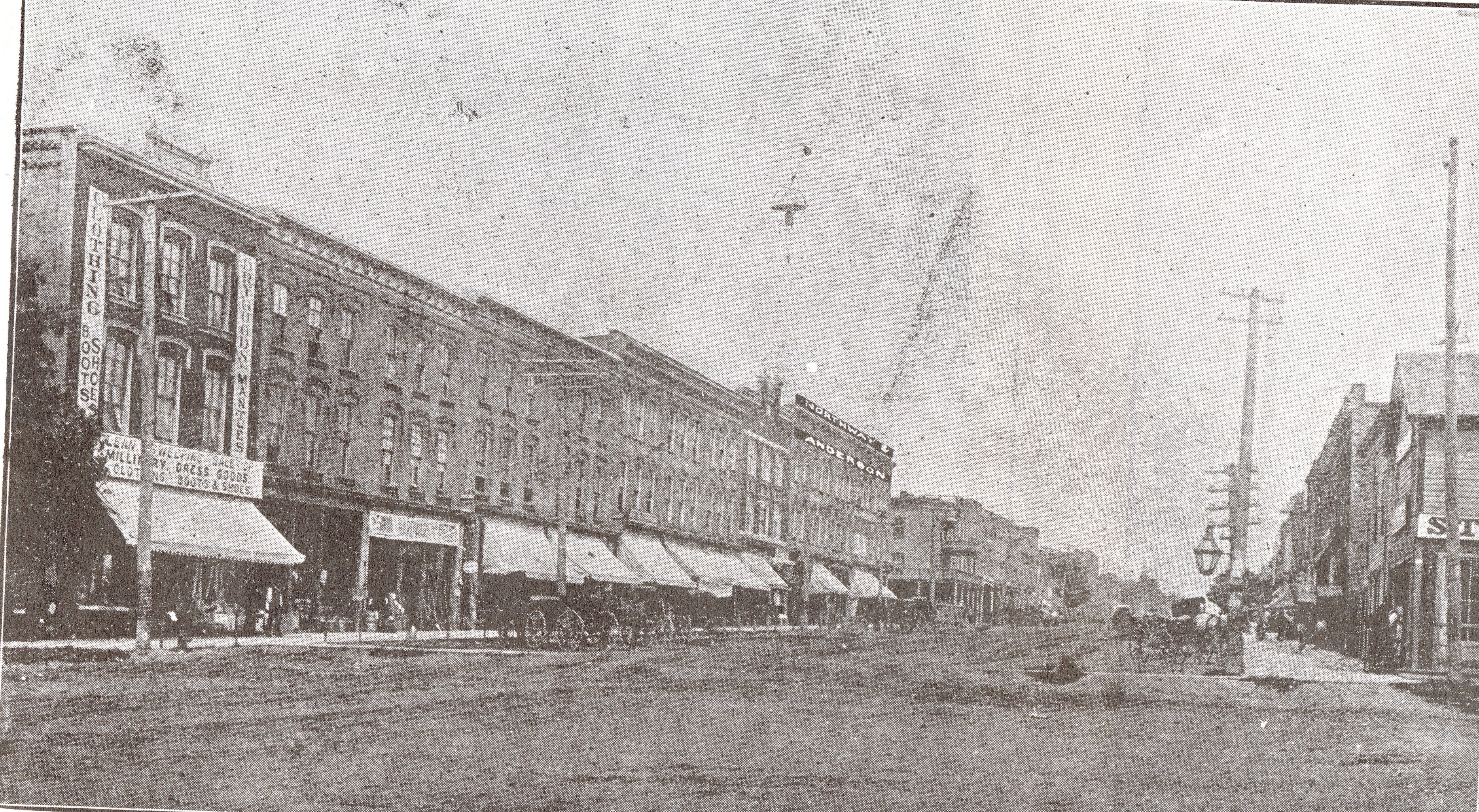 A black and white photograph of Tillsonburg's main street Broadway. The street is dirt and lined with various buildings and businesses on either side. Horse drawn wagons and carriages are parked outside the buildings. Telephone poles also line the street. 
