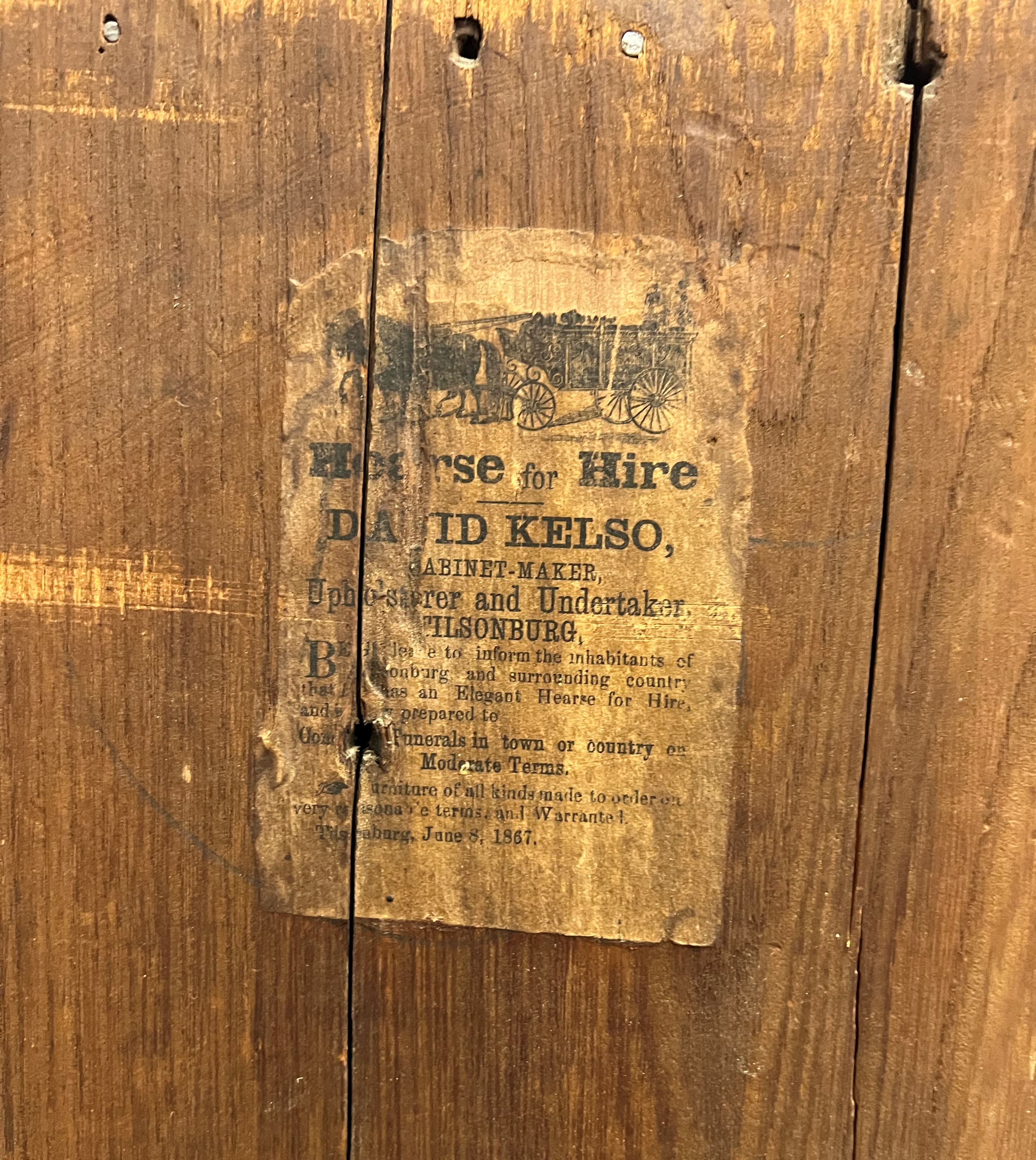 An advertisement pasted onto a wooden board for David Kelso's undertaker business. The advertisement features horses pulling a hearse.