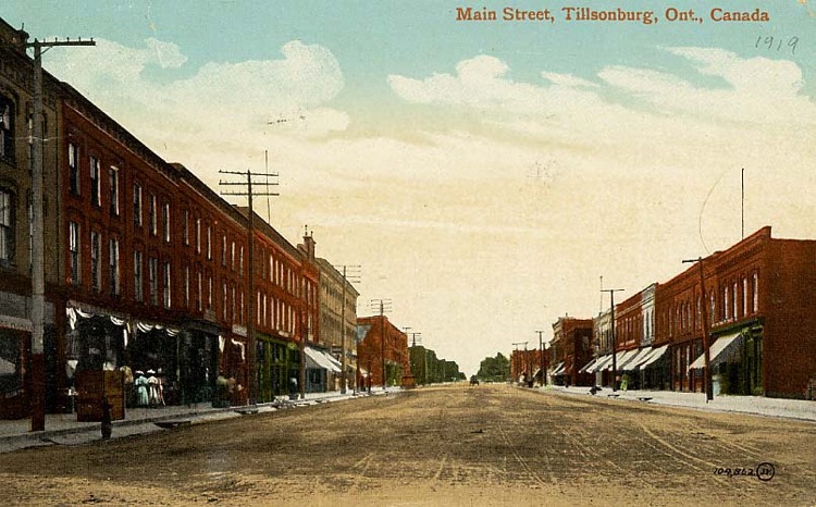 A view of Main Street in Tillsonburg, Ontario. Image is in colour. It is a dirt street line by red and brown brick buildings and store fronts. Some of the buildings have awnings and telephone poles out front. Two women wearing pastel coloured dresses are looking into a store window. 