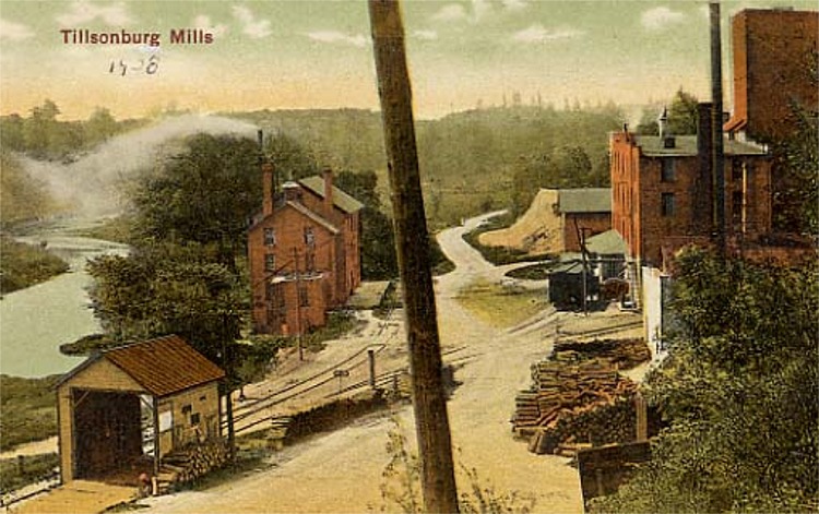 A postcard featuring a red brick mill situated between a dirt road and a creek, smoke is escaping from the smokestack. A large red brick building is across the road with piles of logs and sawdust in front of it.