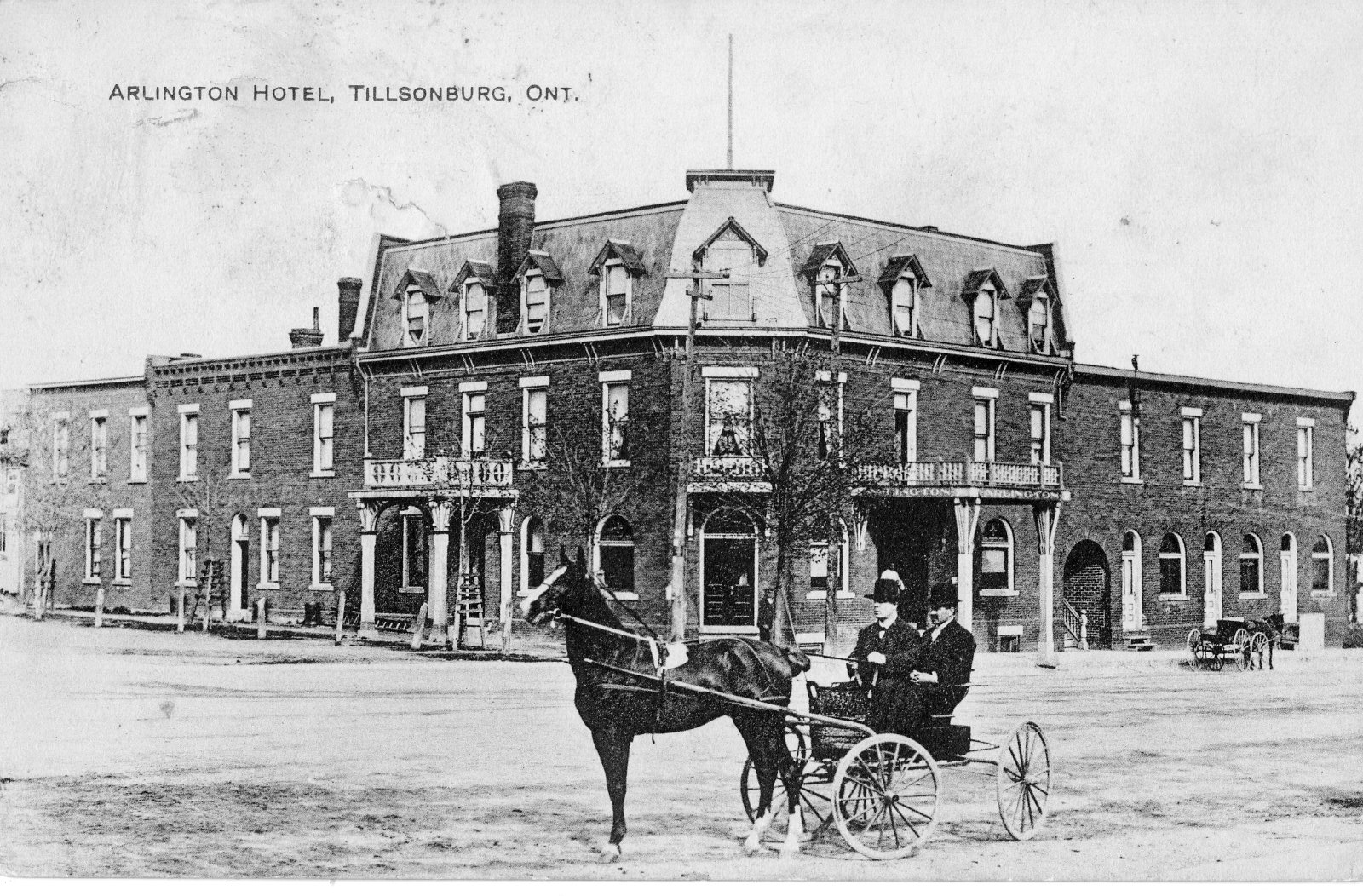 Postcard featuring the Arlington Hotel in Tillsonburg. Two men in a horse-drawn buggy are sitting outside of the hotel in the foreground. Arlington Hotel is a large brick building with a with a mansard style roof.